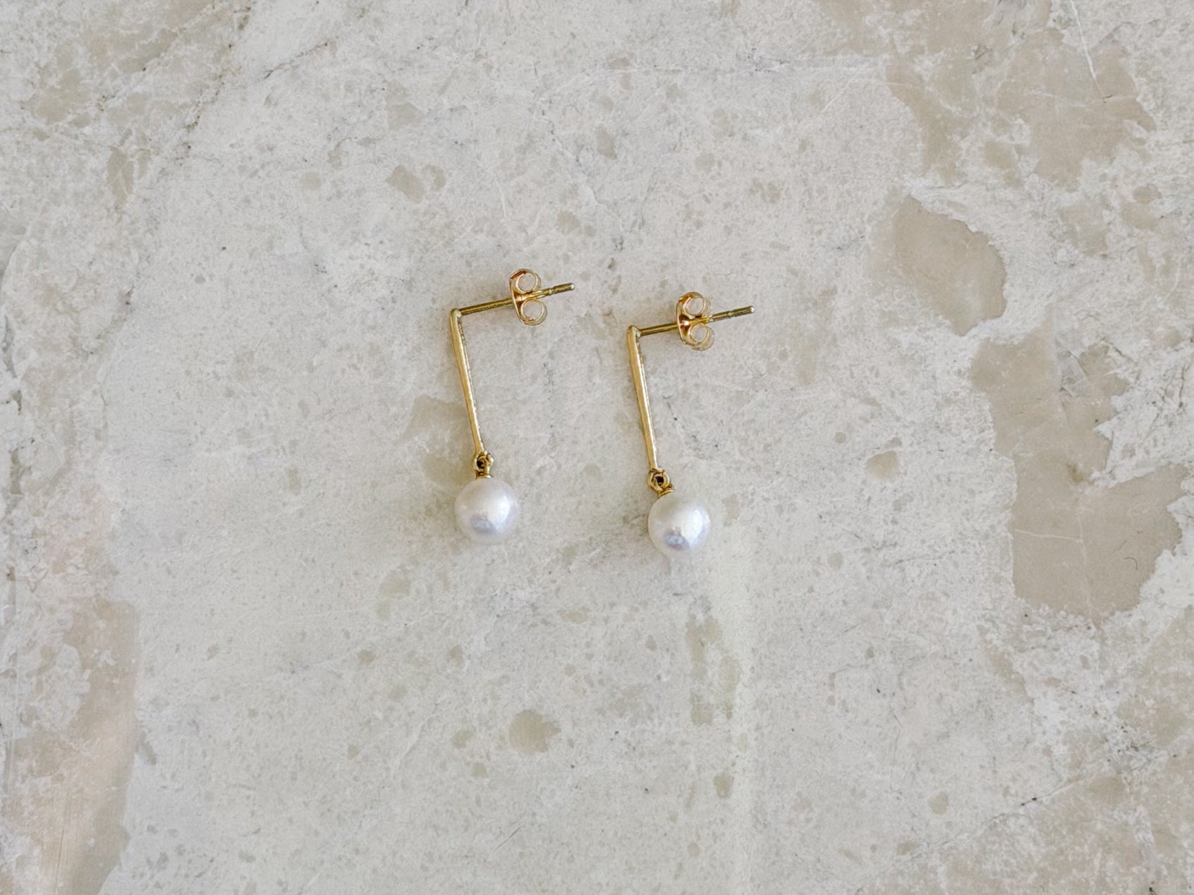 A pair of gold-toned geometric drop earrings with a small white pearl hanging from each, displayed on a light beige stone surface.