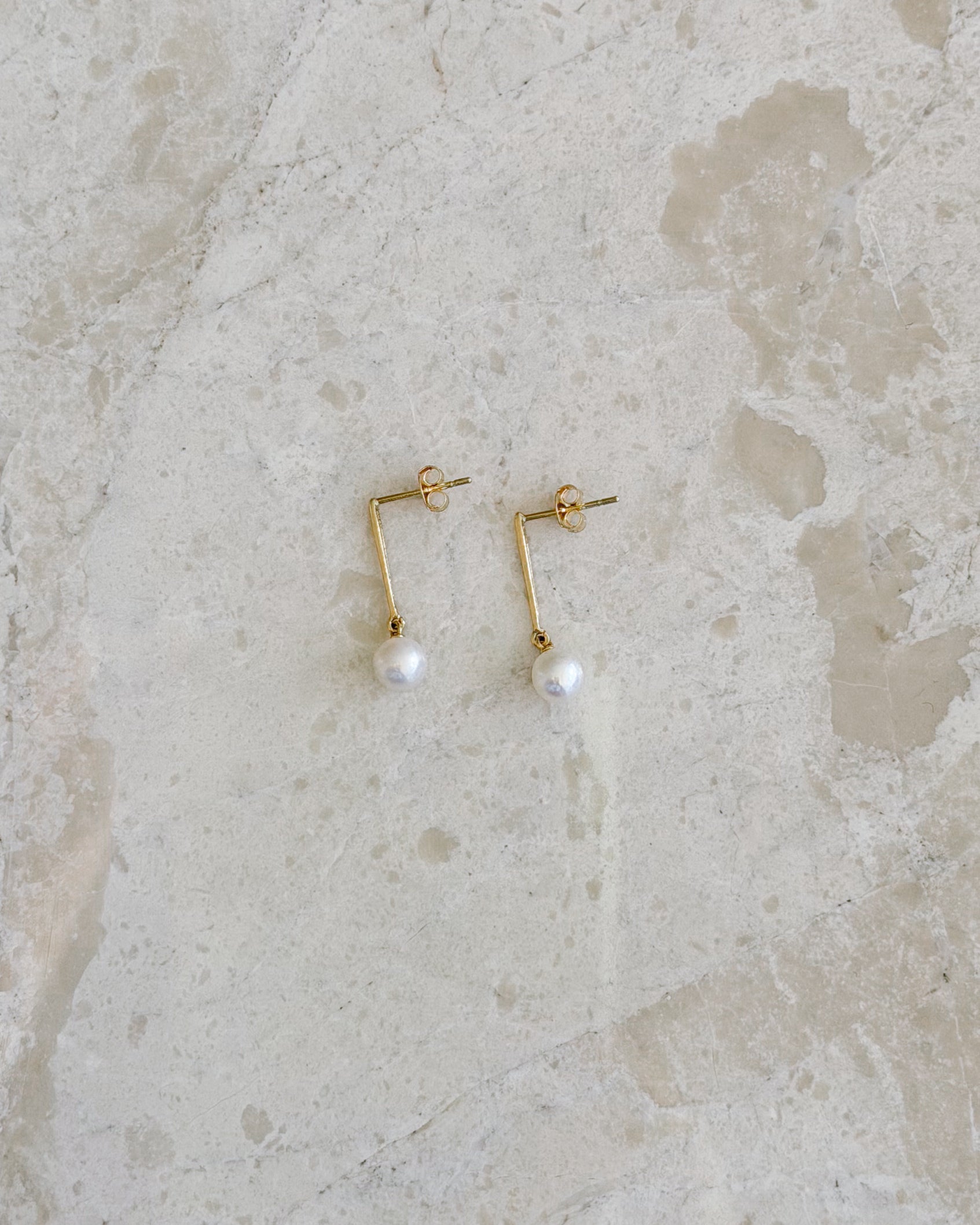 A pair of gold-toned geometric drop earrings with a small white pearl hanging from each, displayed on a light beige stone surface.