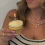 Woman holding a cupcake with a lit candle while wearing a striped sweater and layered gold necklaces.