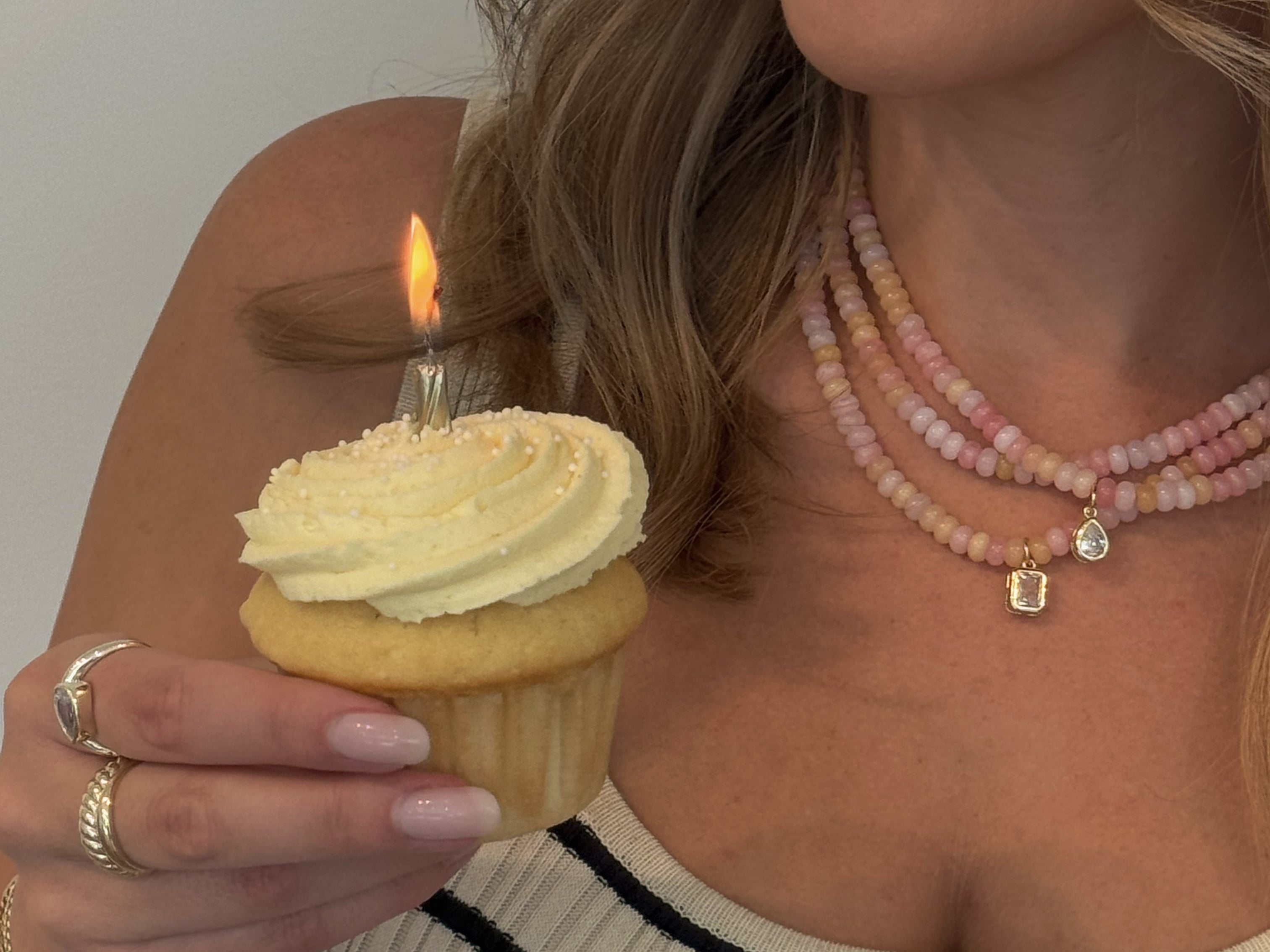 Woman holding a cupcake with a lit candle while wearing a striped sweater and layered gold necklaces.